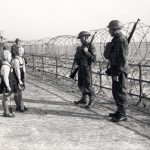 Photo of soldiers by the seaside railings, which is protected with barbed wire. Two children are speaking with them as they are guarding the perimeter.
