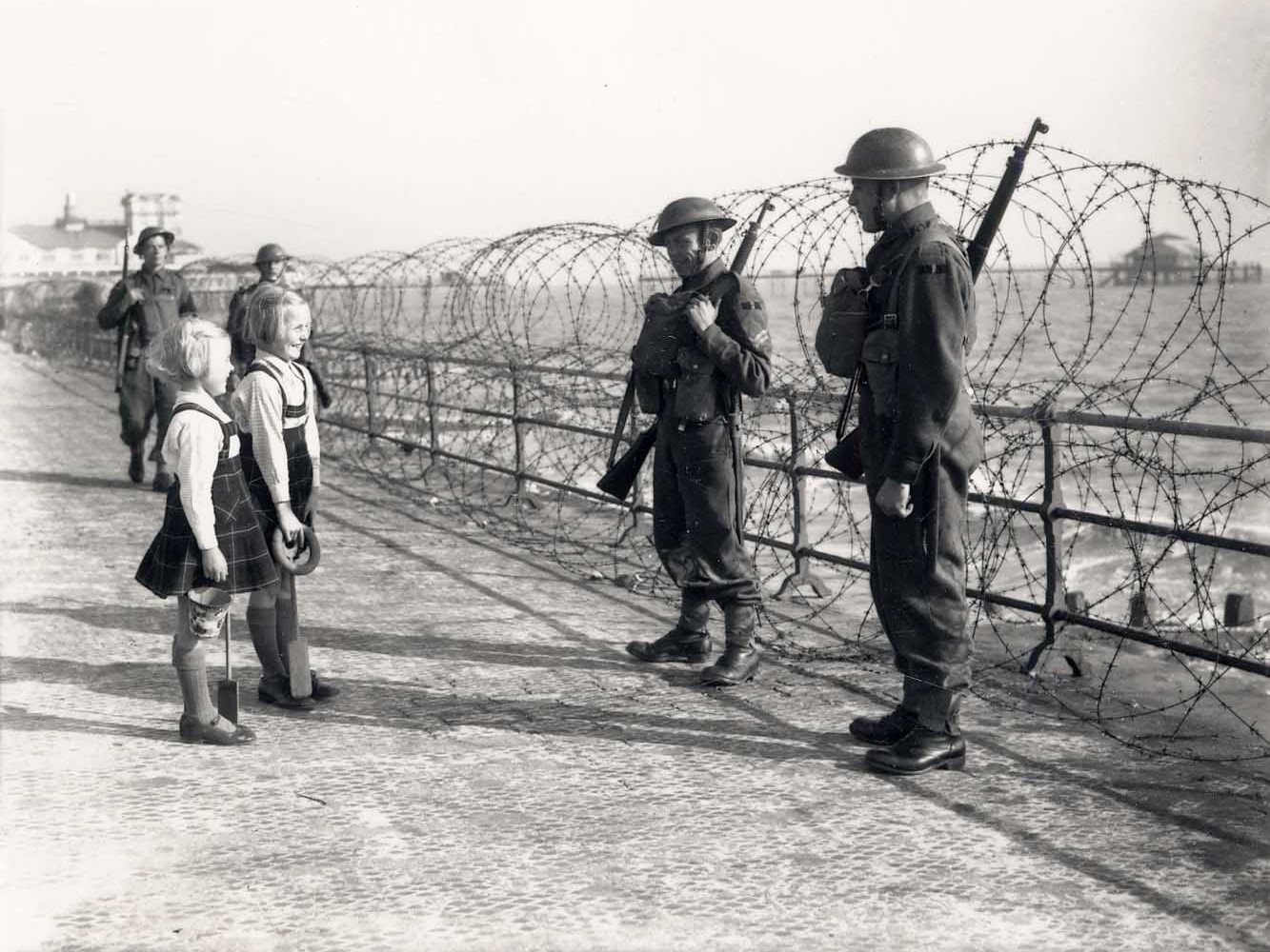 Photo of soldiers by the seaside railings, which is protected with barbed wire. Two children are speaking with them as they are guarding the perimeter.